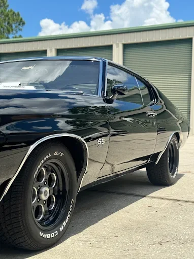 Black vintage Chevrolet Chevelle SS muscle car parked on concrete driveway with a green building and blue sky in the background.