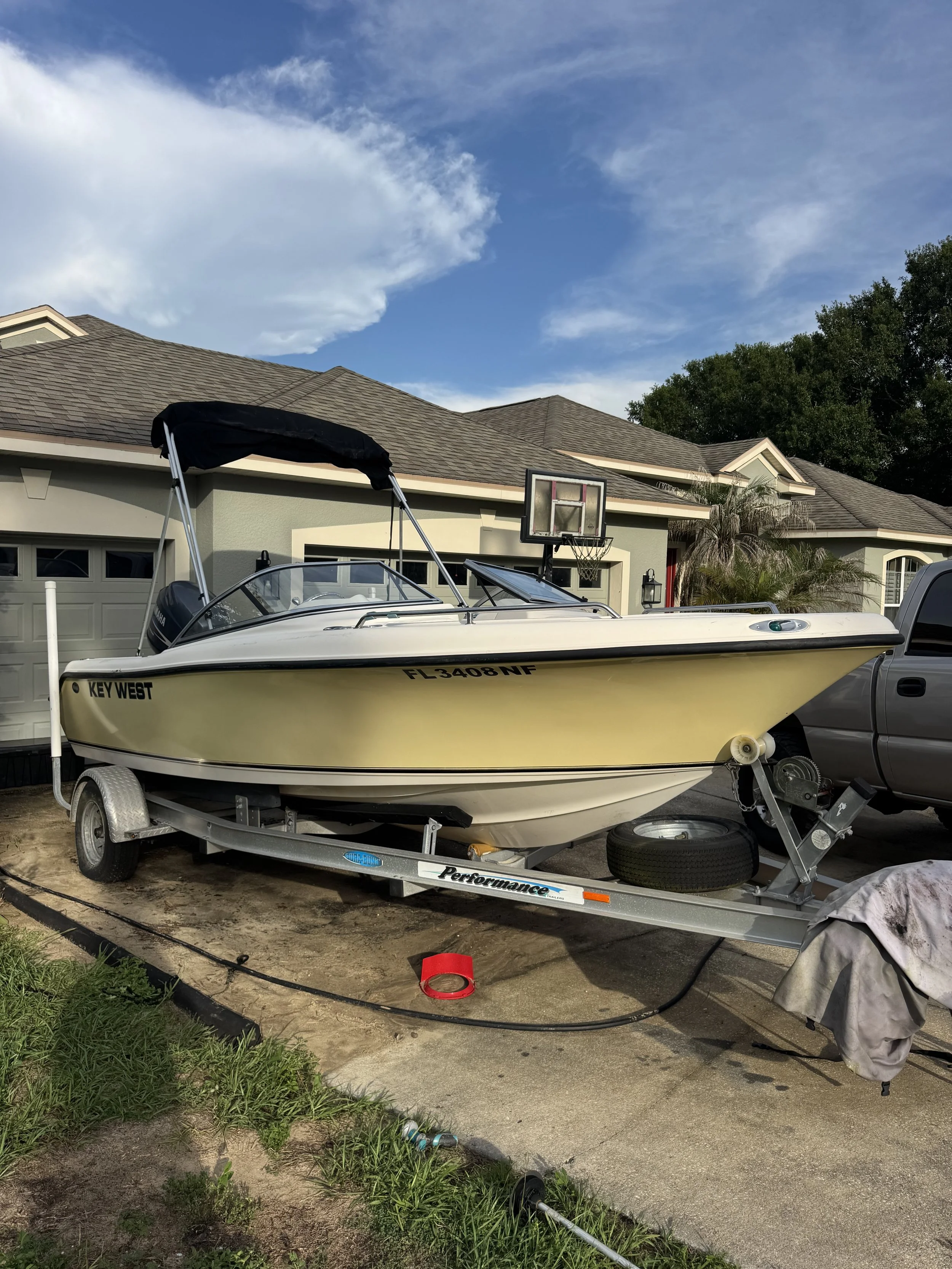 A yellow speedboat on a trailer parked in a driveway in front of a house, with a basketball hoop and trees in the background.