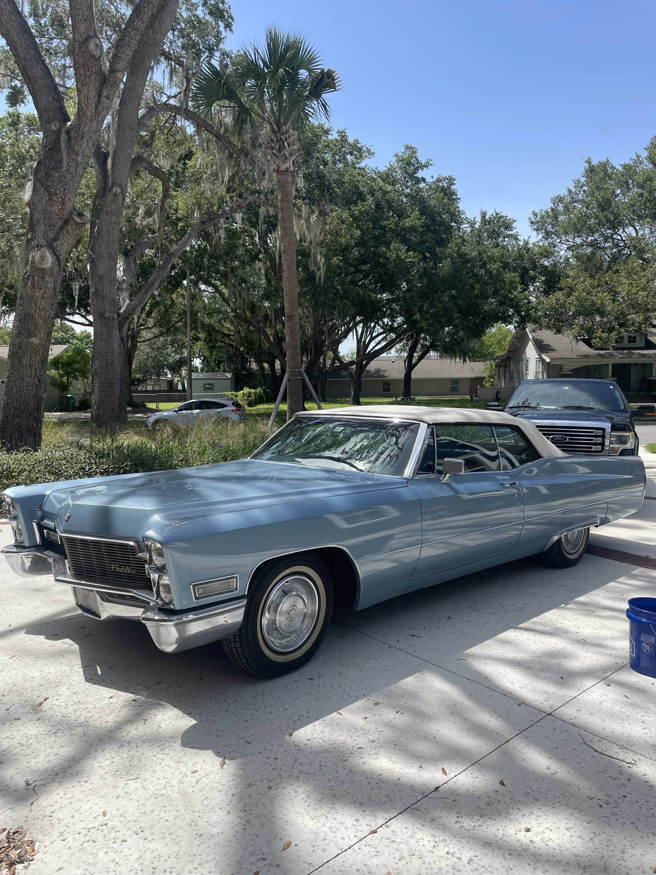 A vintage light blue Cadillac convertible parked on a concrete driveway, with trees, houses, and cars in the background under a clear blue sky.
