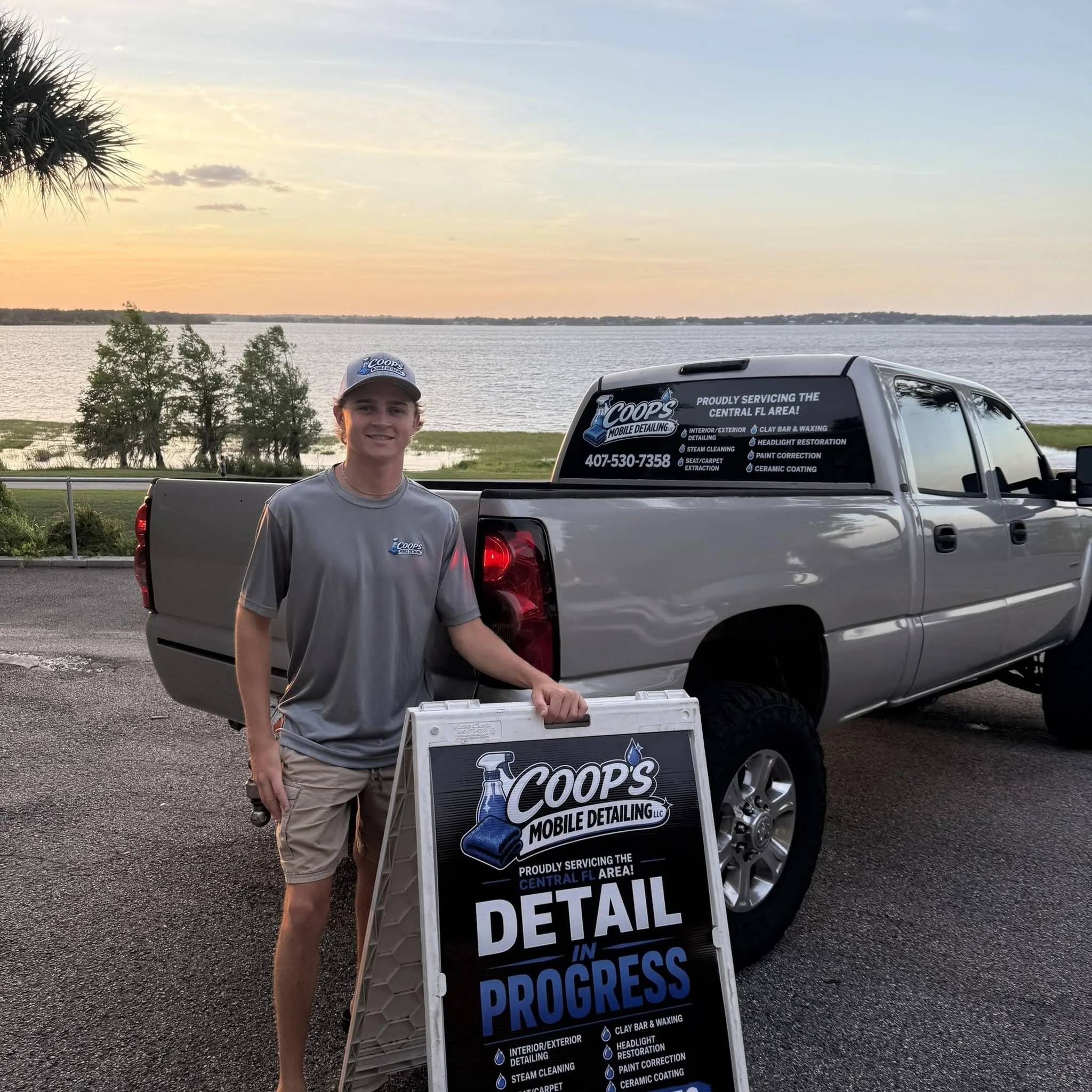 Young man standing next to a vehicle with a sign that reads 'Coop's Mobile Detailing, LLC' and 'In Progress' under a sunset sky, near a body of water and trees.