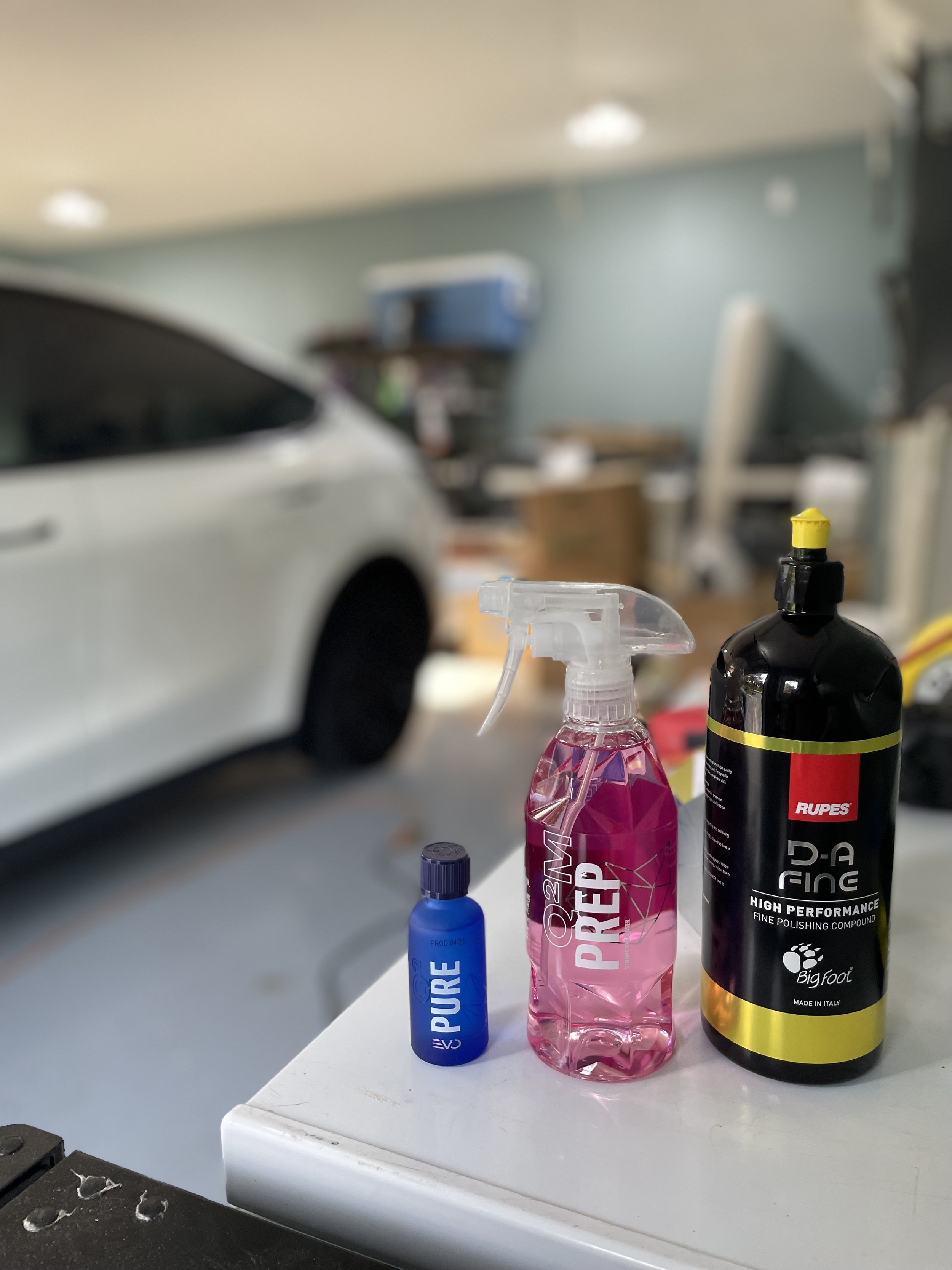 Various car care bottles on a white surface in a garage, with a car in the background.