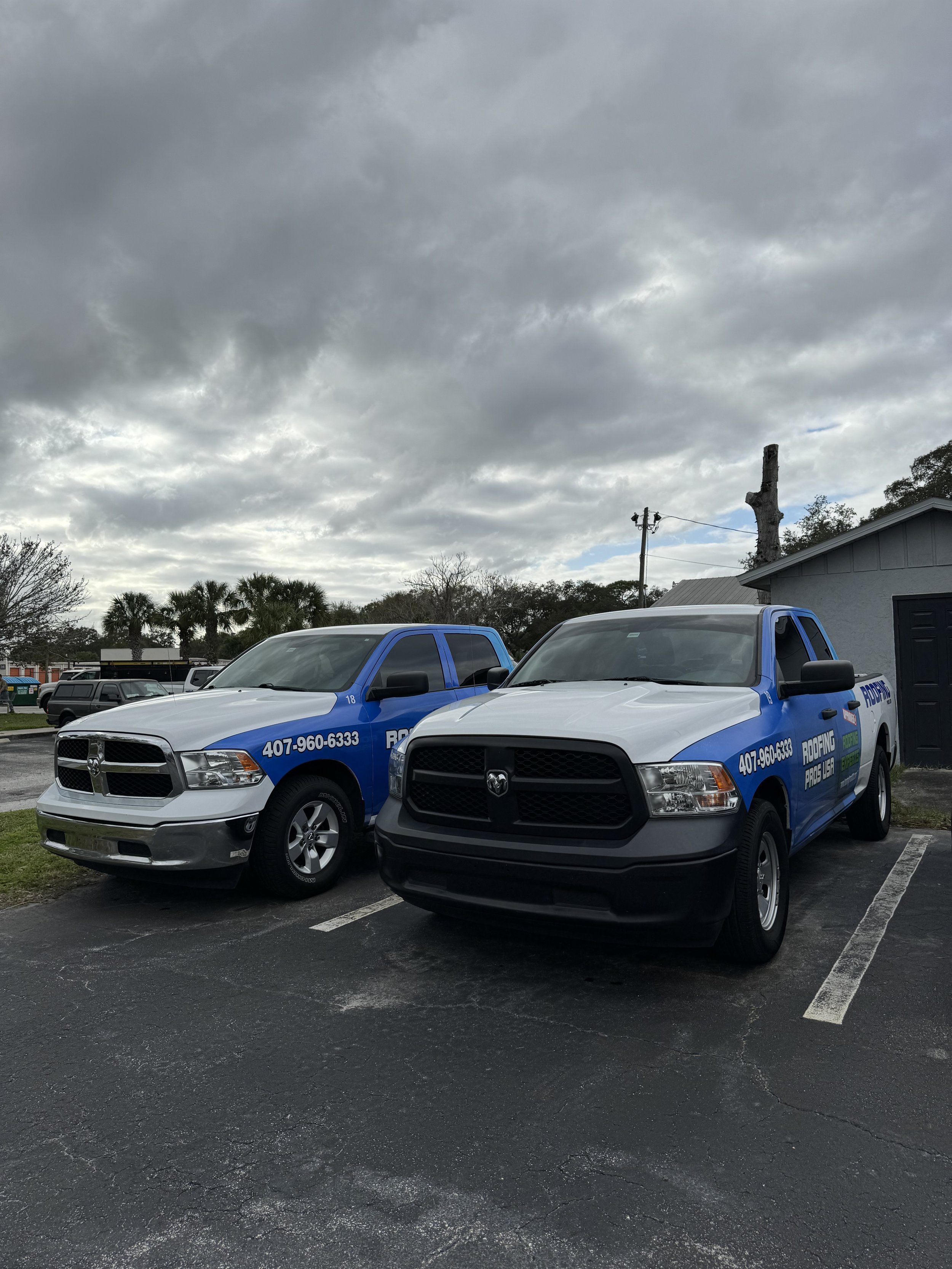 Two pickup trucks parked in a parking lot under a cloudy sky; both trucks have blue and white graphics, advertisements for roofing services, with contact information and slogan visible.