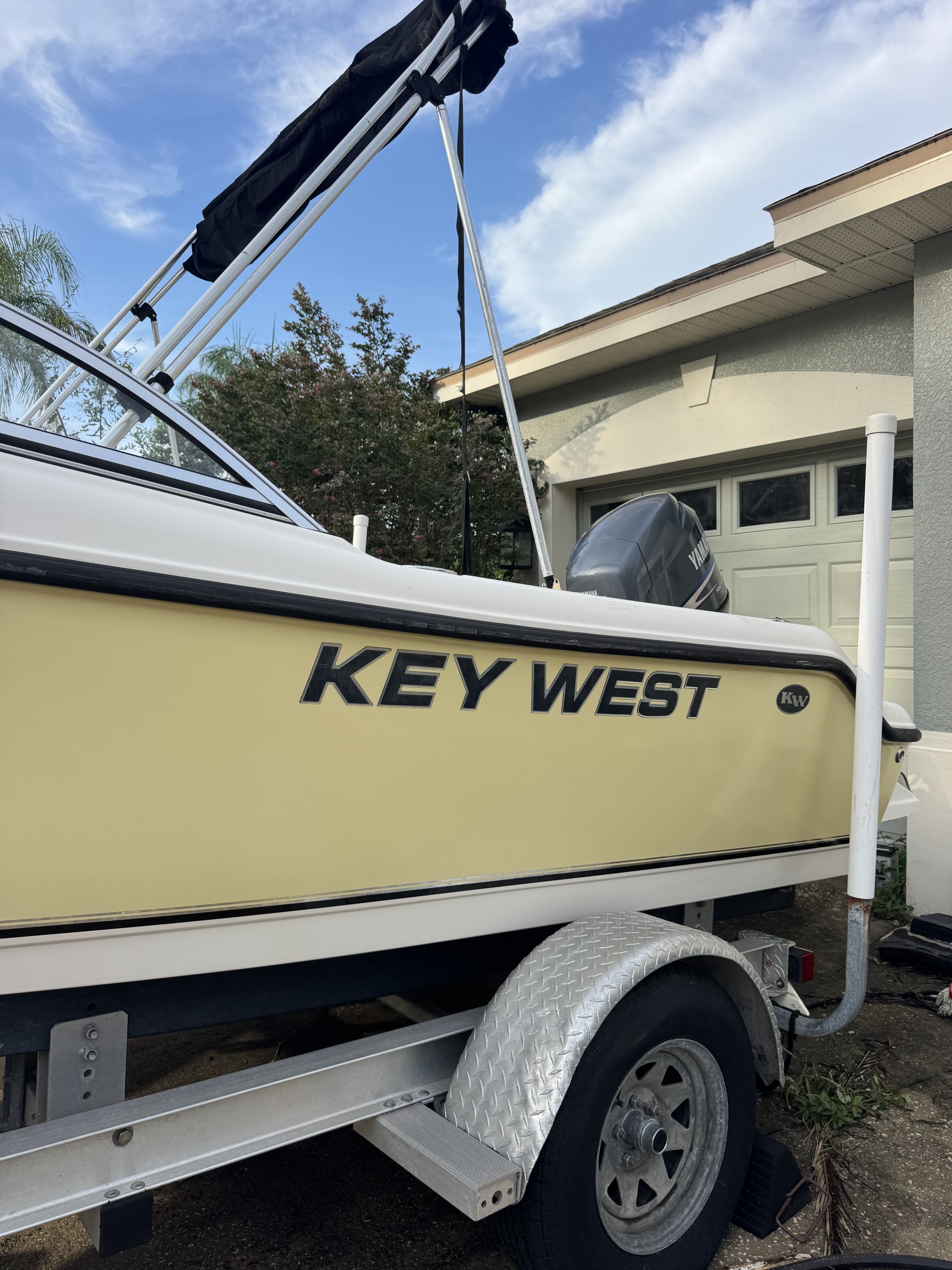 A boat on a trailer parked in a driveway with a house and trees in the background. The boat has a Yamaha outboard motor and a canopy or cover folded back.