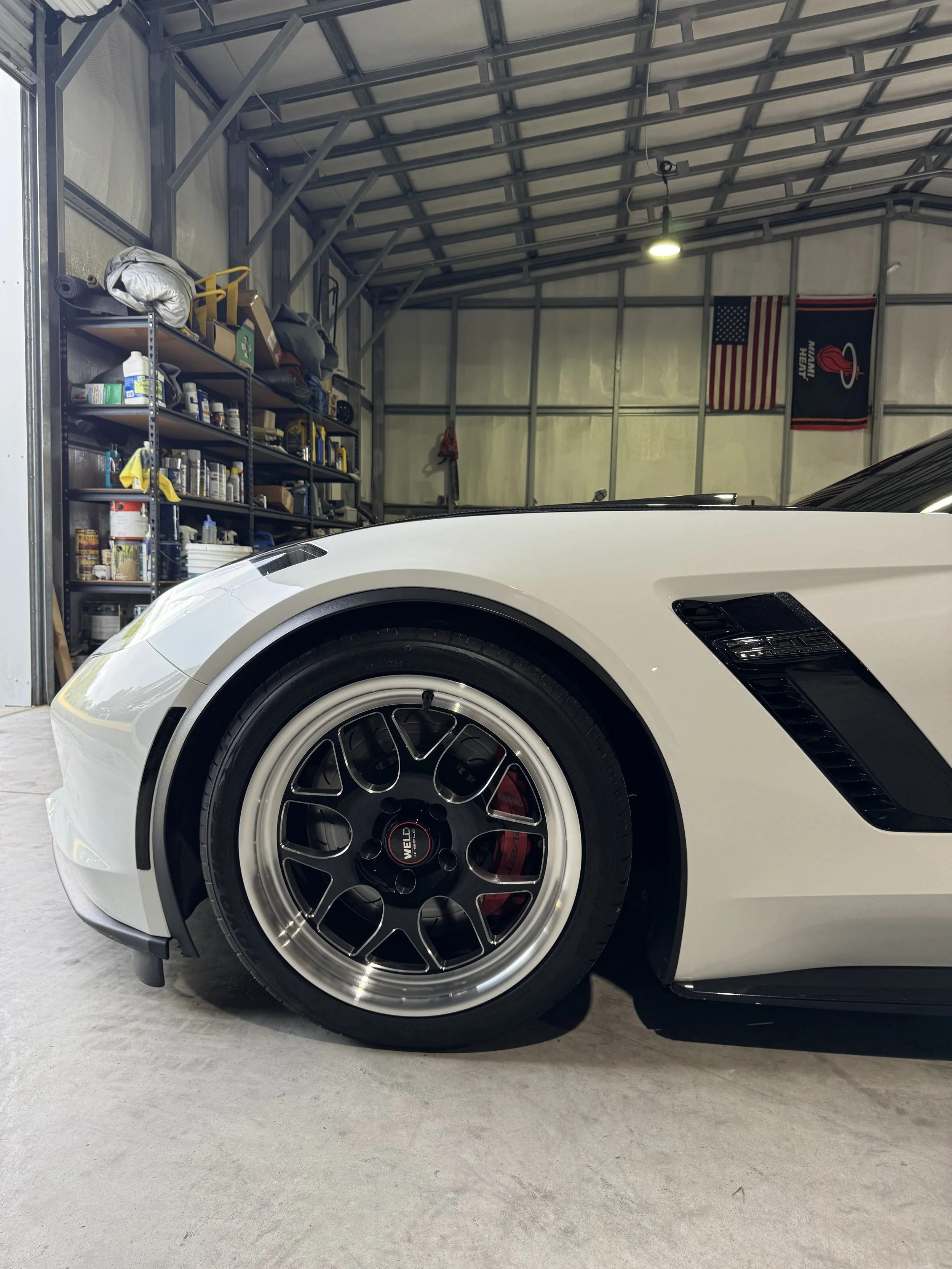 Close-up of a white sports car with a black wheel, parked inside a garage with metal shelves and banners on the wall.