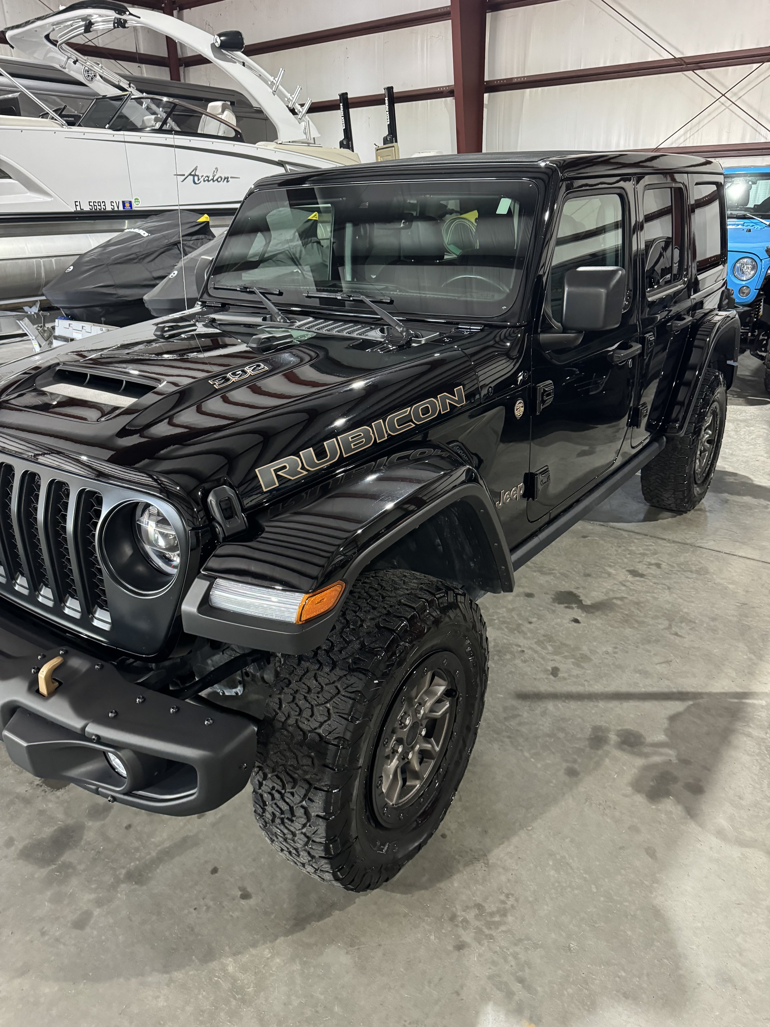 A black Jeep Rubicon with large off-road tires parked inside a warehouse. Behind it, a white boat and a blue vehicle are visible.