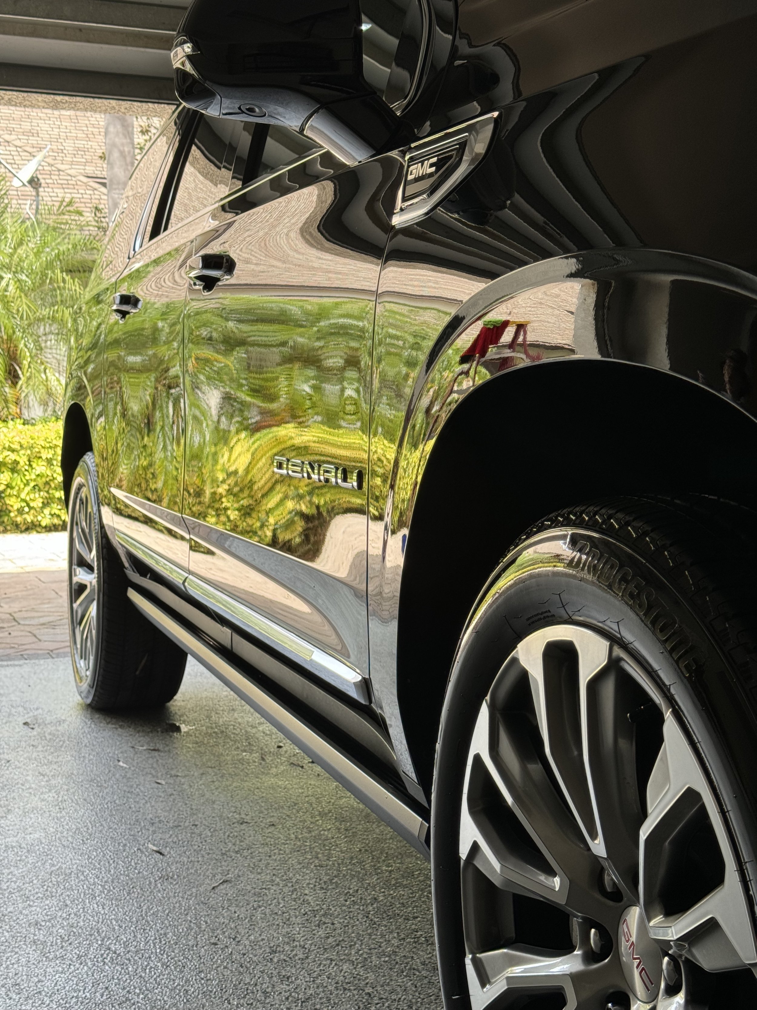 A black GMC vehicle with shiny, reflective paint parked outdoors near green shrubbery and a building. The image shows the side profile of the vehicle, focusing on the rear door and wheel.
