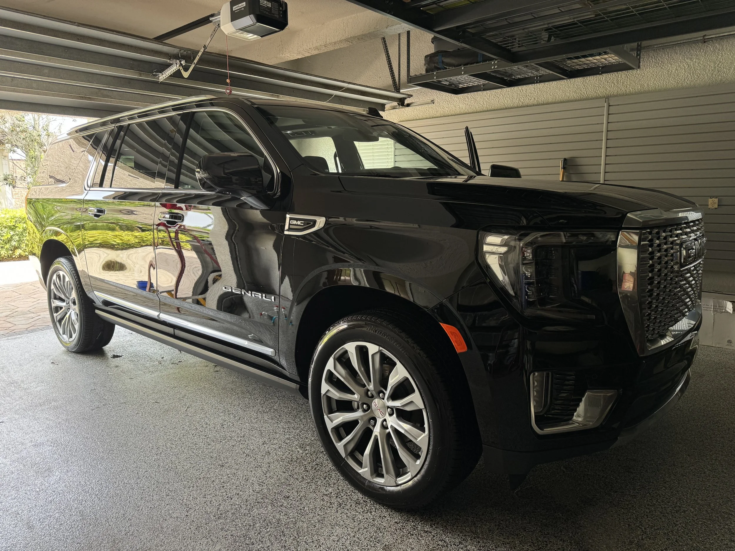 A black GMC Denali SUV parked in a garage with a gray textured floor, with the garage door partially open showing some trees and sky outside. The vehicle has shiny alloy wheels and tinted windows.