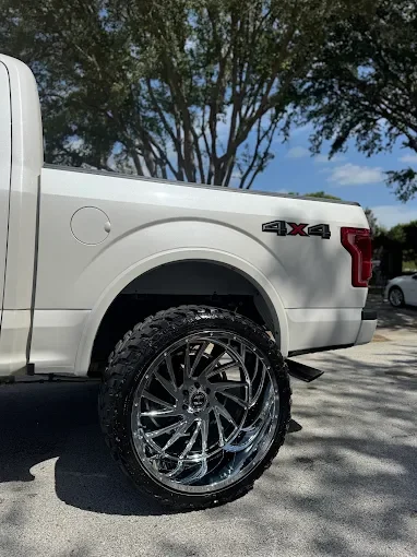 Close-up of the rear side of a white pickup truck with chrome wheels and a two-tone black and gray tire, parked outdoors under a tree.