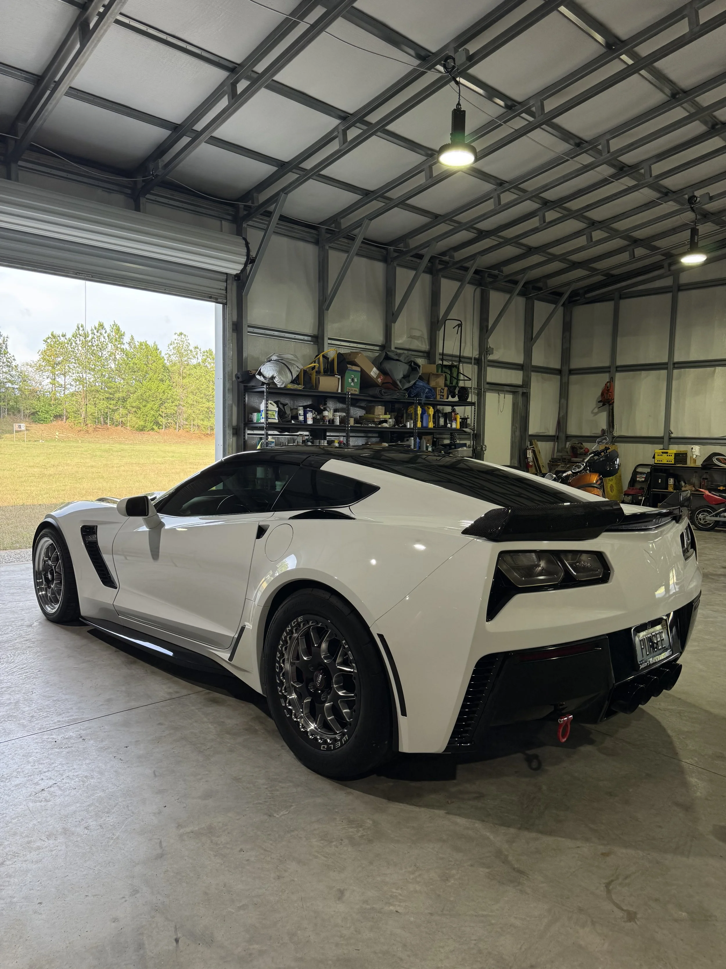 A white sports car with black accents parked inside a garage with a metal frame and tools on shelves.