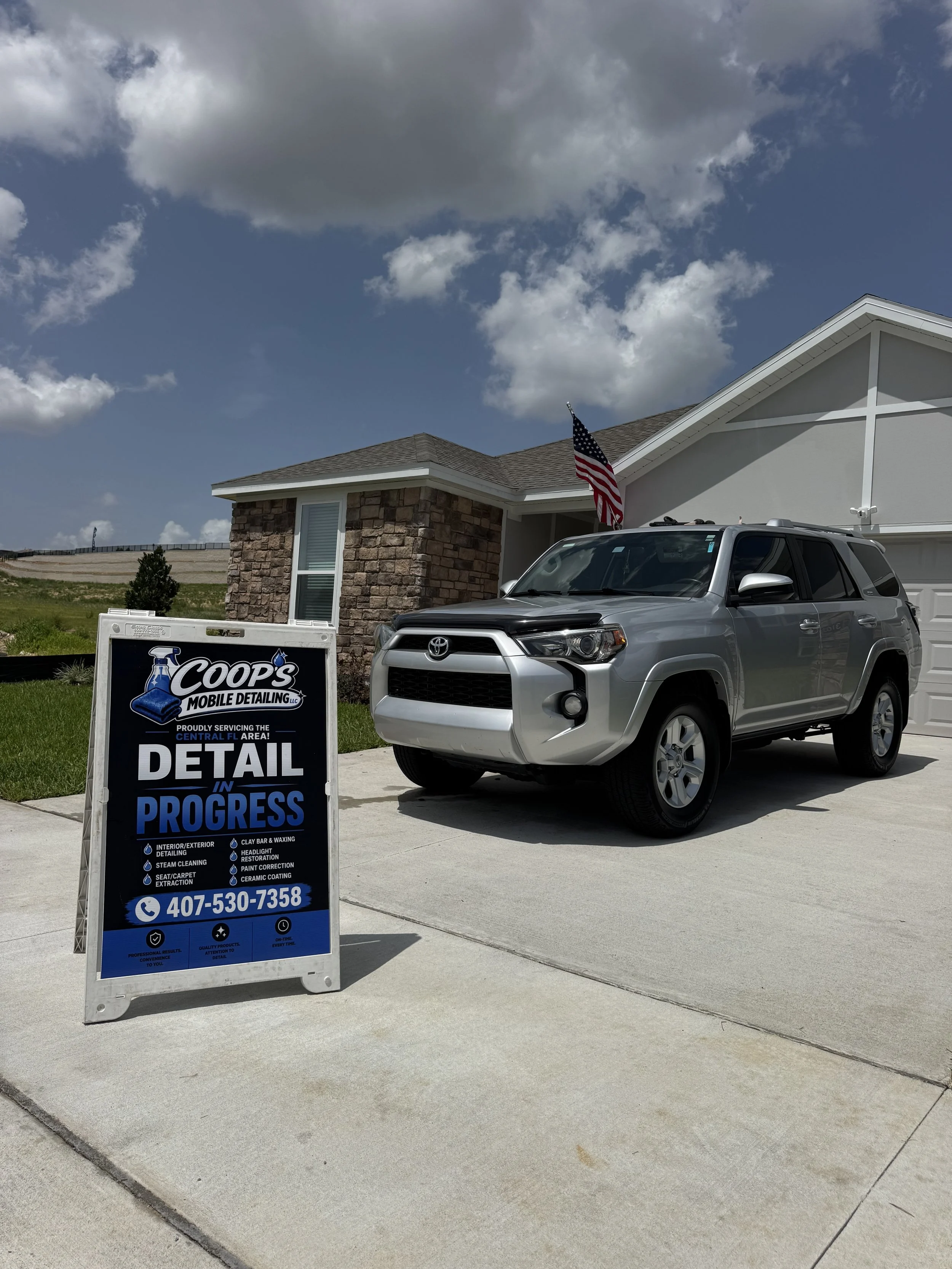 Signboard for Coops Mobile Detailing located on a concrete driveway in front of a house with a gray Toyota SUV and an American flag.