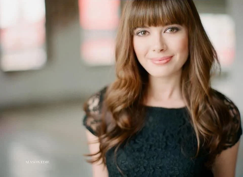 A woman with long, wavy red hair and bangs, smiling and looking at the camera, wearing a black lace top, in a well-lit indoor setting with blurred windows in the background.
