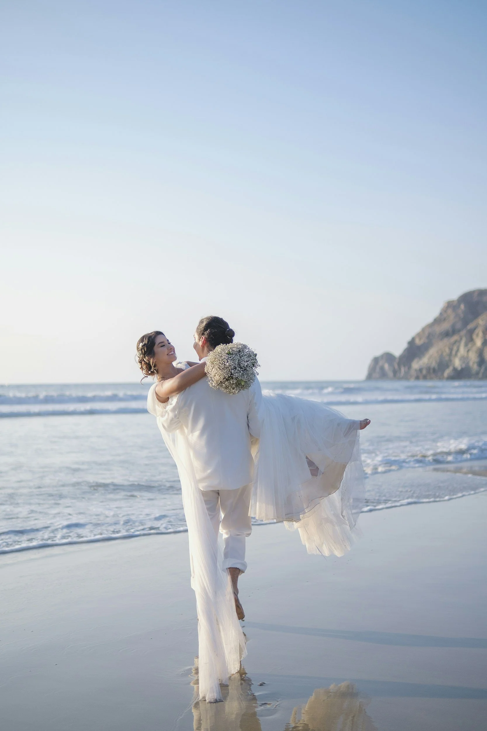 A couple dressed in wedding attire, the man is holding the woman in his arms on a beach, with the ocean and cliffs in the background. The woman is holding a bouquet of flowers and both are smiling.