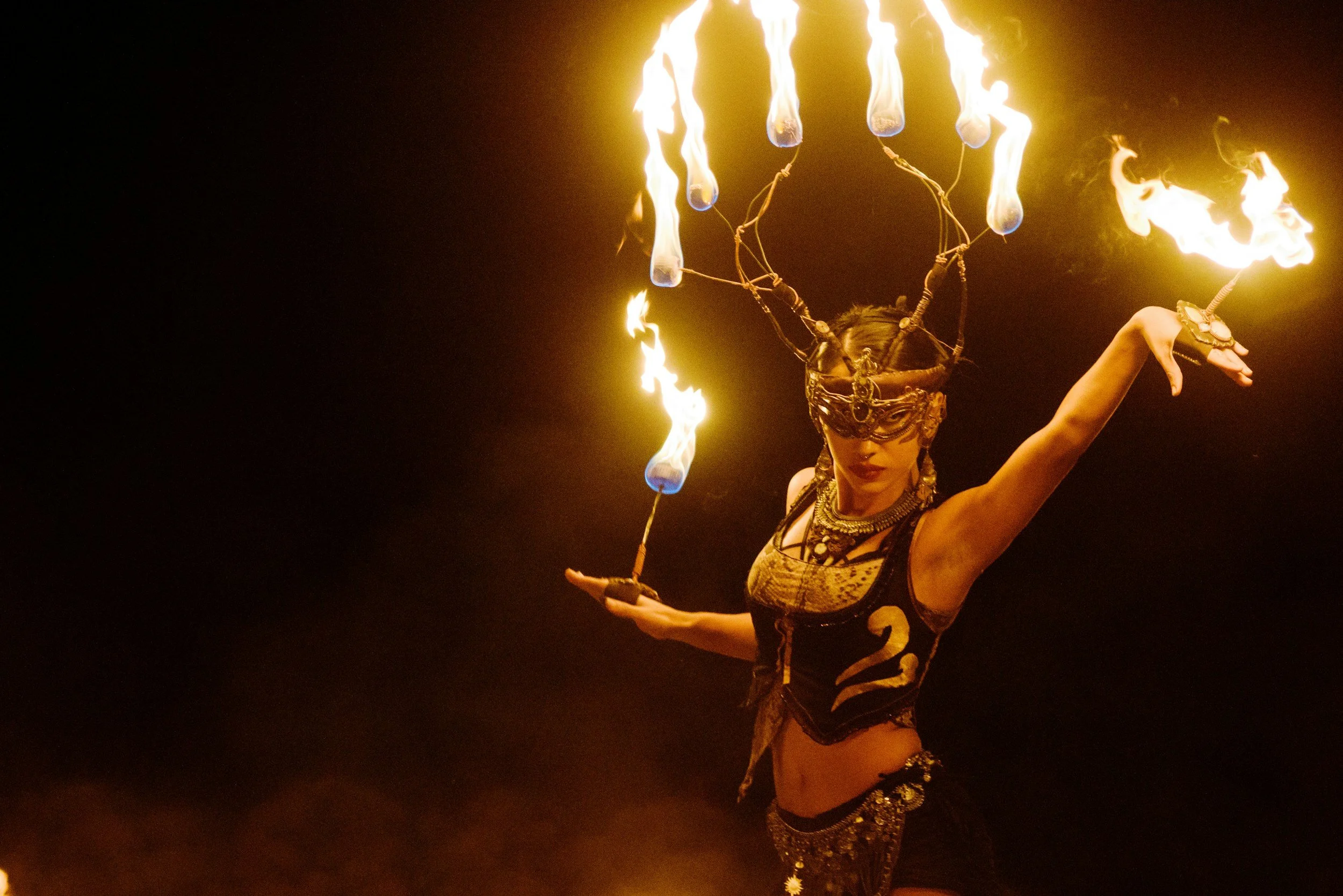 A female performer dressed in elaborate, traditional costume with decorative jewelry, performing a fire dance at night, with multiple flames burning from fire torches.