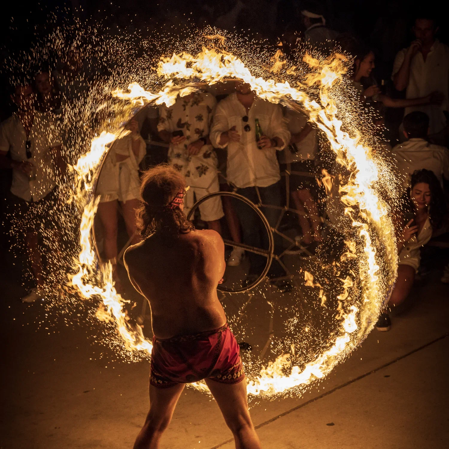 A fire performer twirling a flaming fire hoop creates a ring of fire around their body, with spectators watching at night.