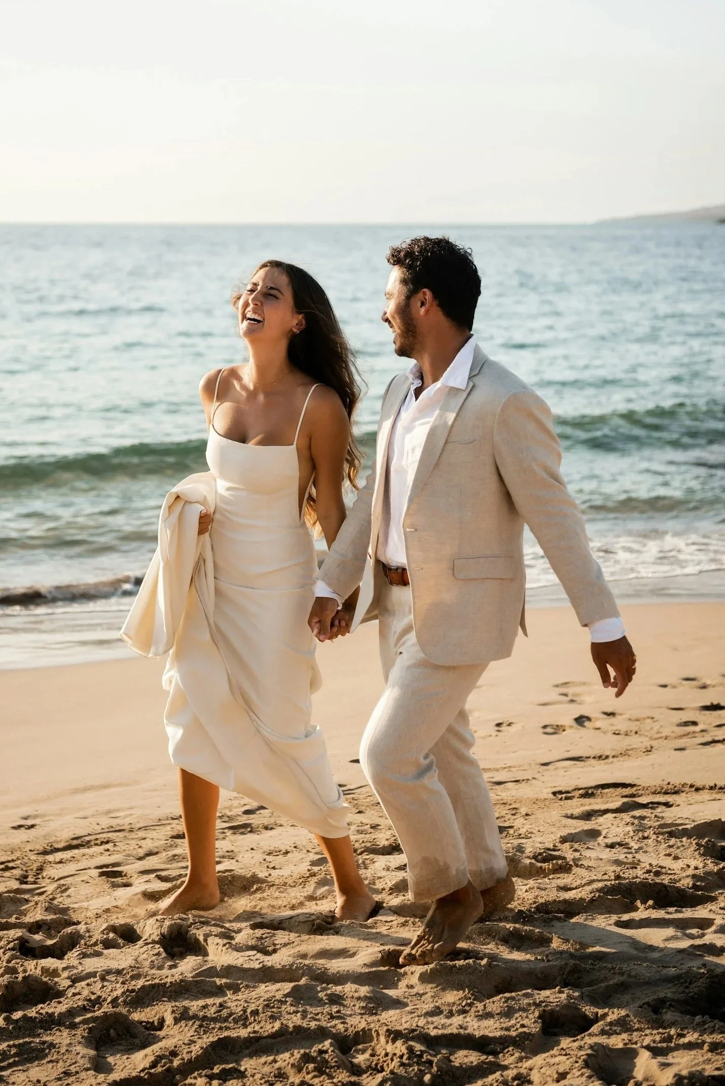 A couple dressed in wedding attire walking on a sandy beach, holding hands and laughing with the ocean in the background, during daylight.