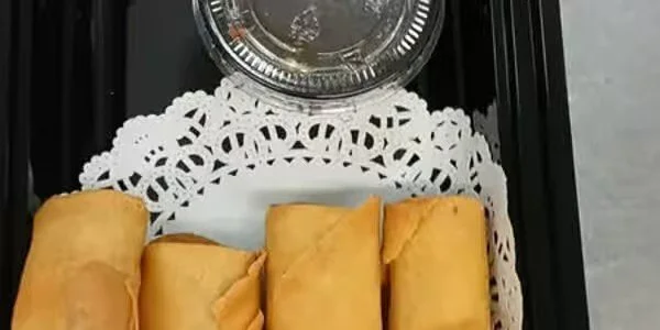 Four egg rolls on a tray with a decorative paper liner, a container of dipping sauce above them, and a small cup of sauce next to the container.