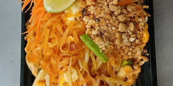 A plate of pad thai noodles topped with crushed peanuts, lime wedge, shredded carrots, and chopped green onions.