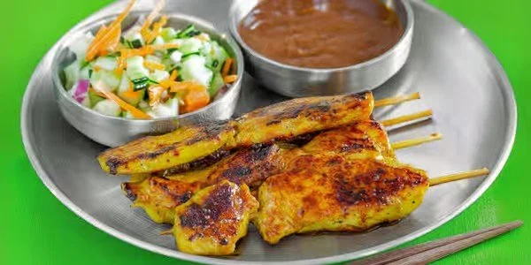 Chicken satay skewers with peanut sauce and vegetable salad on a metal tray.