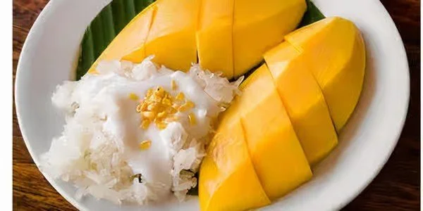 A bowl of fresh mango slices with sticky rice topped with coconut cream and yellow garnish, on a wooden surface.