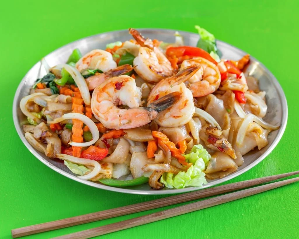 Stir-fried shrimp and vegetables on a silver plate with chopsticks on a green surface.