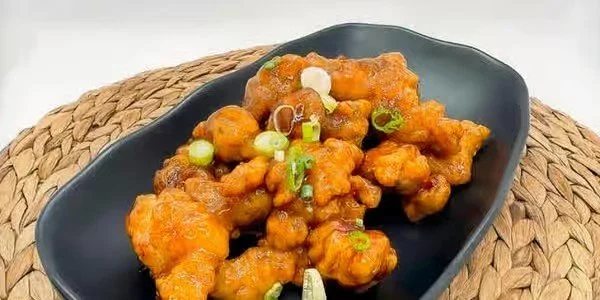 Bowl of fried chicken pieces coated in orange sauce, garnished with chopped green onions, on a woven placemat.
