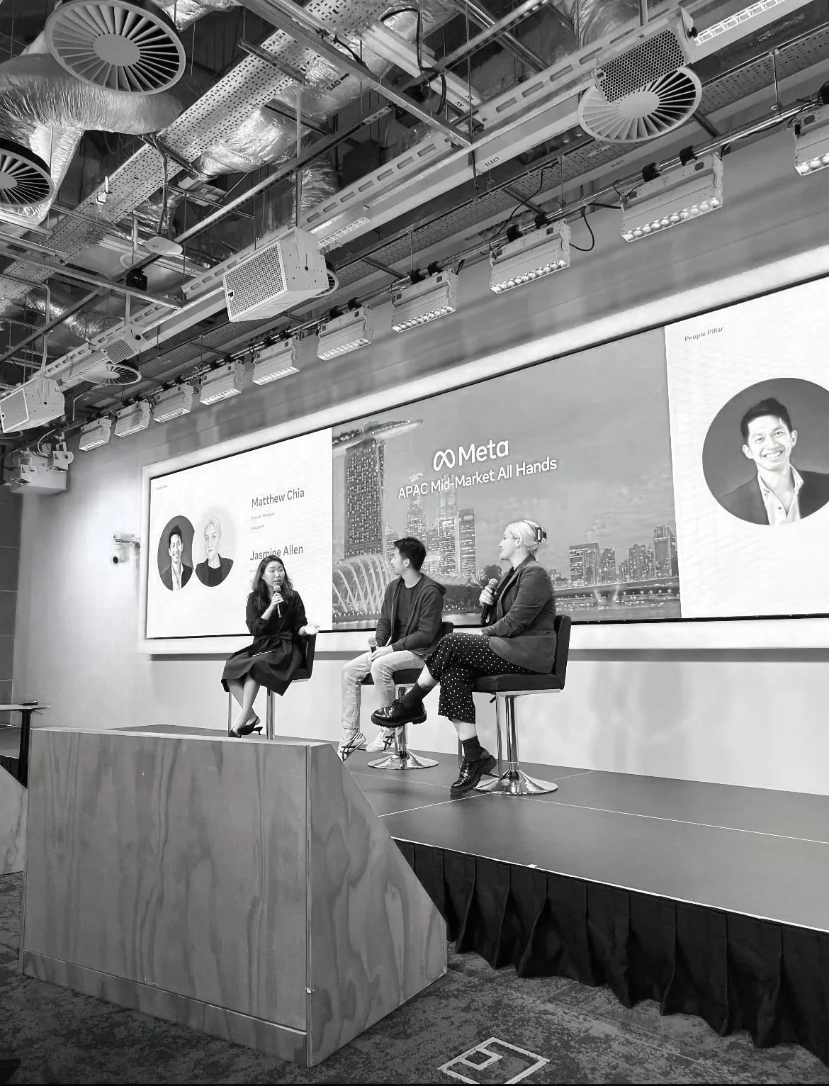A panel discussion taking place in a conference room with three speakers seated on a stage, and a large screen behind them displaying their names and the event details for Meta, APAC Mid-Market. The room has an industrial ceiling with exposed pipes and ventilation.