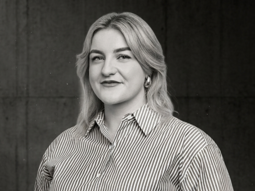 Black and white portrait of a woman with shoulder-length hair, wearing a striped collared shirt, smiling slightly, standing in front of a plain dark background.