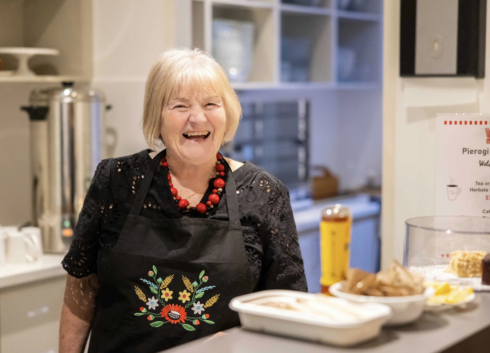 Smiling volunteer in an embroidered apron serving at a food stall at a Dom Polski event.