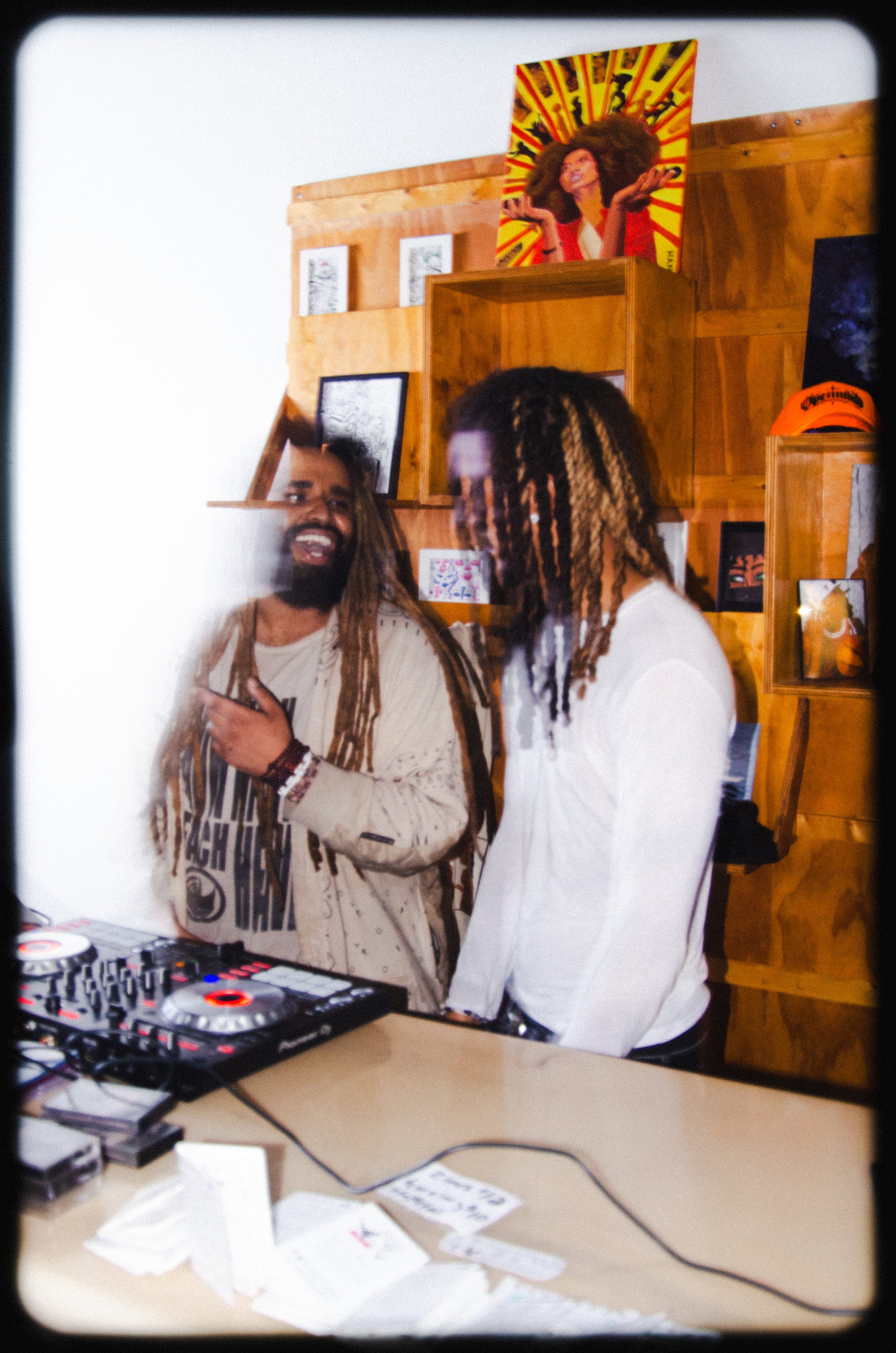 Two men with dreadlocks, one with a beard, standing near DJ equipment and engaged in conversation, in a room with wooden wall shelves and artwork, with a portrait of a woman on the top shelf.