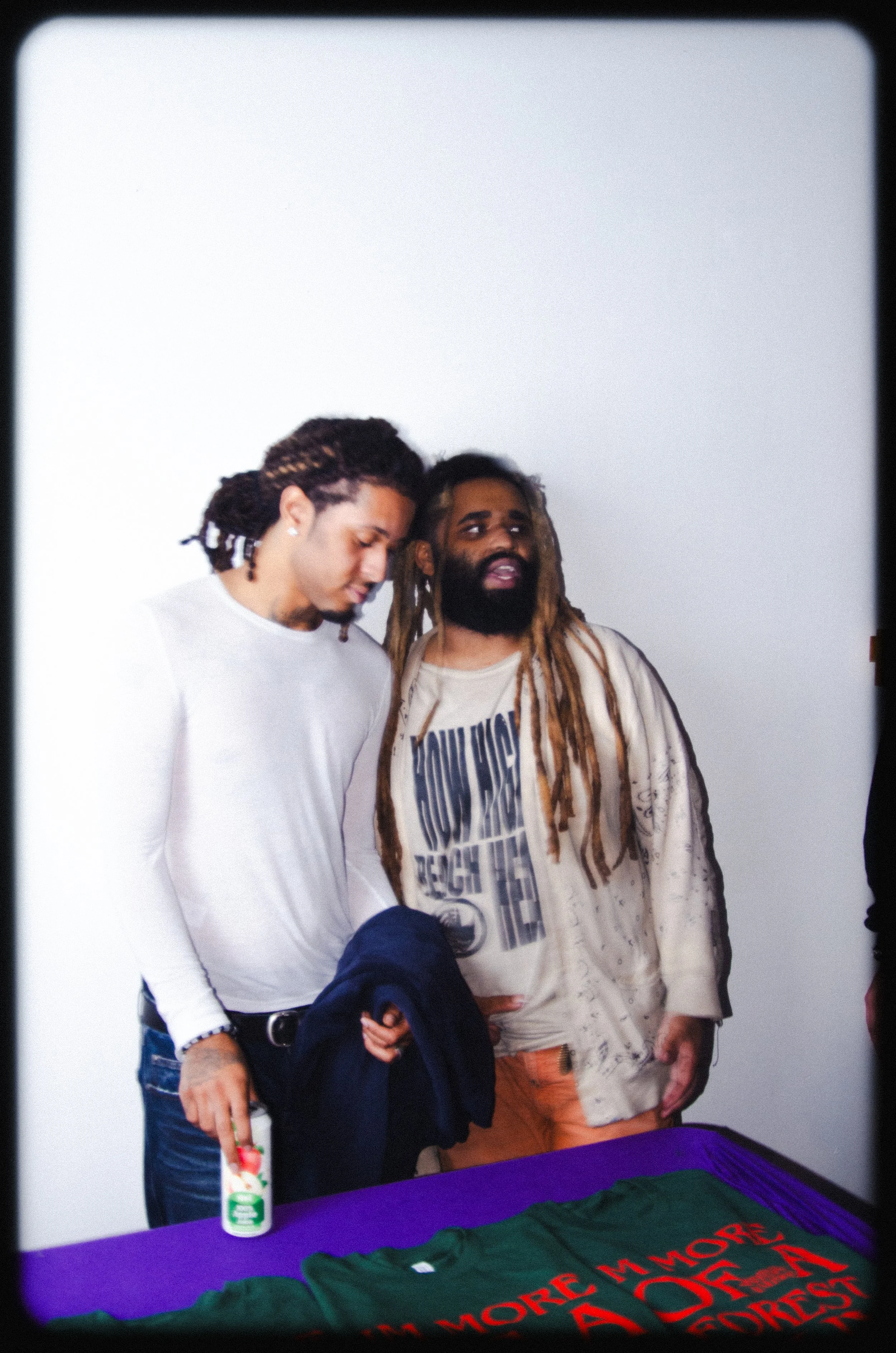 Two men with dreadlocks standing next to each other in front of a white wall, one wearing a white long-sleeve shirt and the other wearing a beige shirt with text, with a purple table in front of them covered with green cloths and red text.