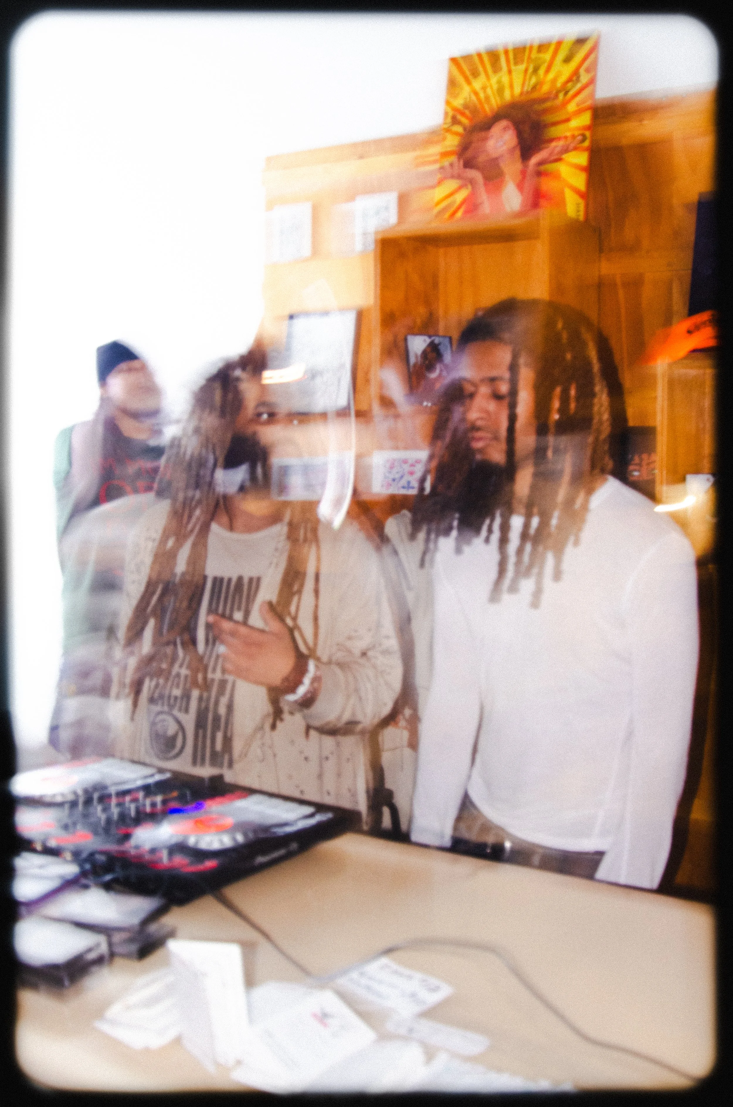A desk with a computer monitor, a DJ controller, and scattered papers. Behind the desk, three young people are visible; one holding a phone, another with braided hair, and the third with glasses and a mask.