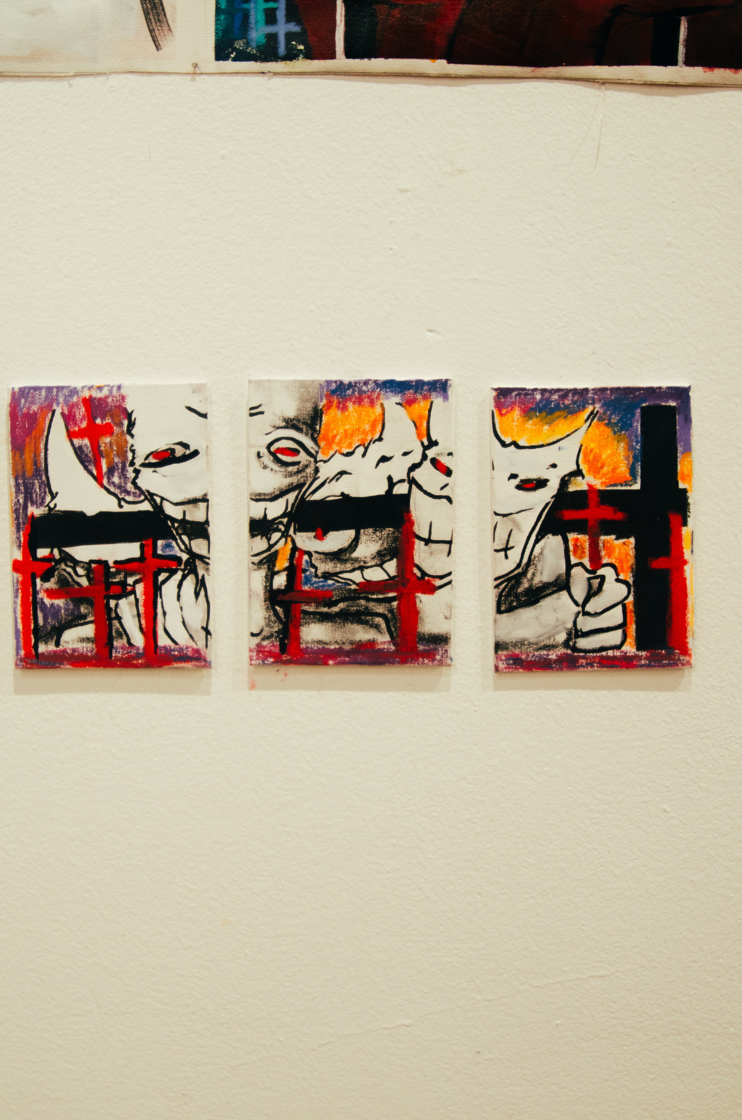 Three small, colorful paintings of a clown with red eyes, orange hair, and a wide smile, displayed on a light-colored wall.