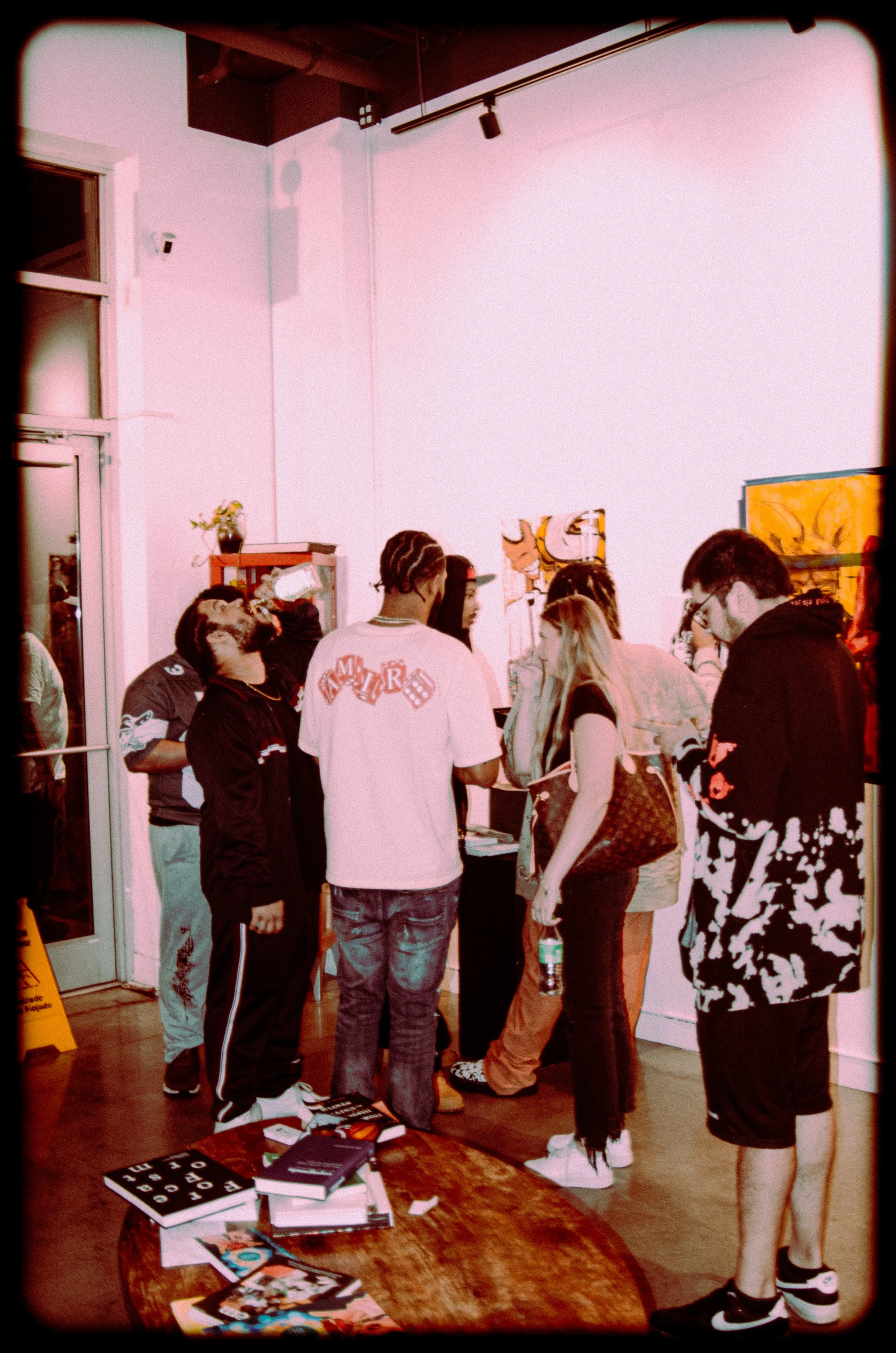 A group of young people gathers around a table with books and magazines in an art gallery or bookstore. Some are looking at the items on the table while others are talking or looking at artwork on the walls.