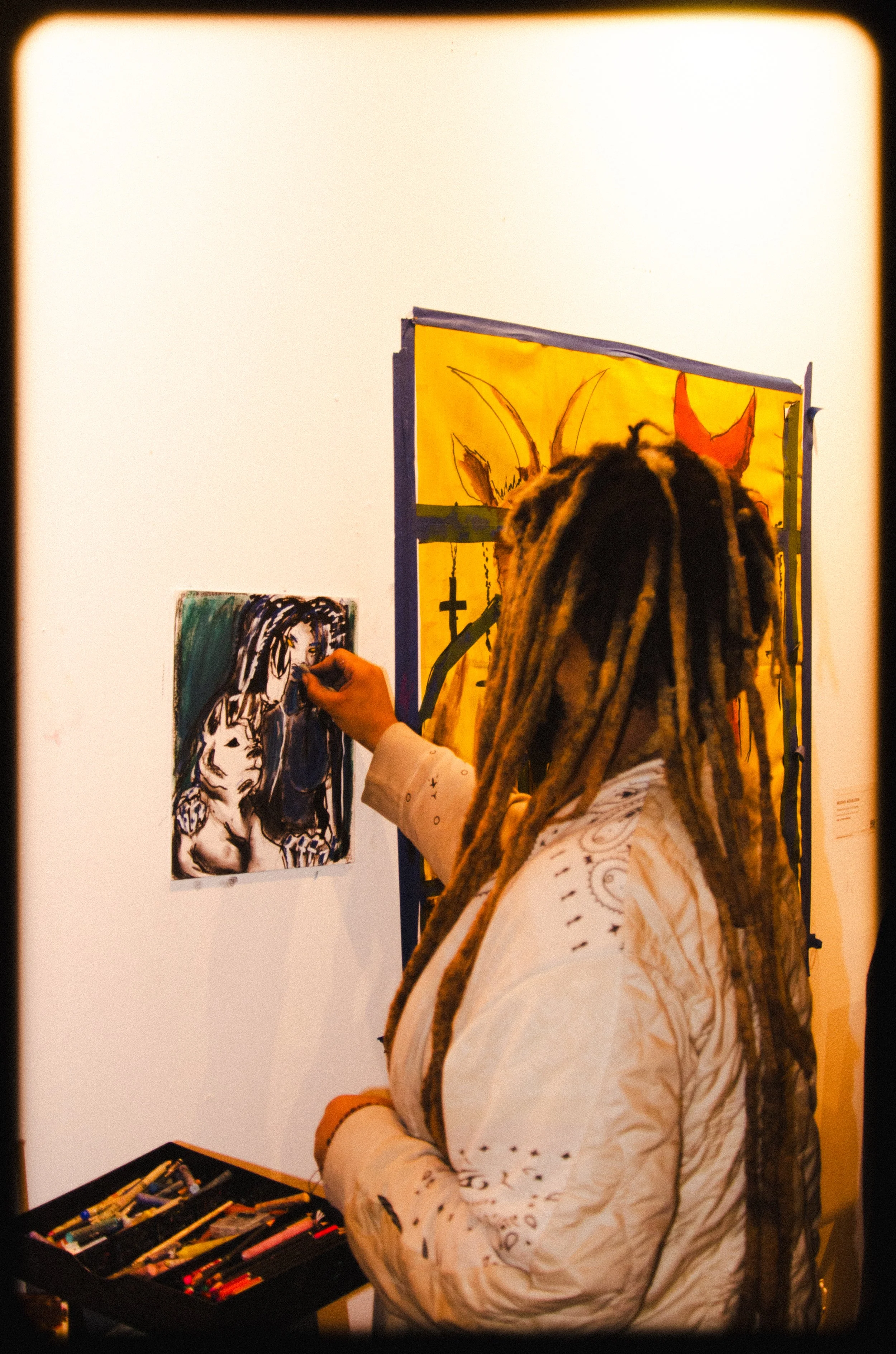 A person with dreadlocks wearing a white long sleeve shirt is drawing on a small canvas with black and white stick figure art. They are standing next to a display of colorful art supplies in a black box, with artwork visible on the wall behind them.