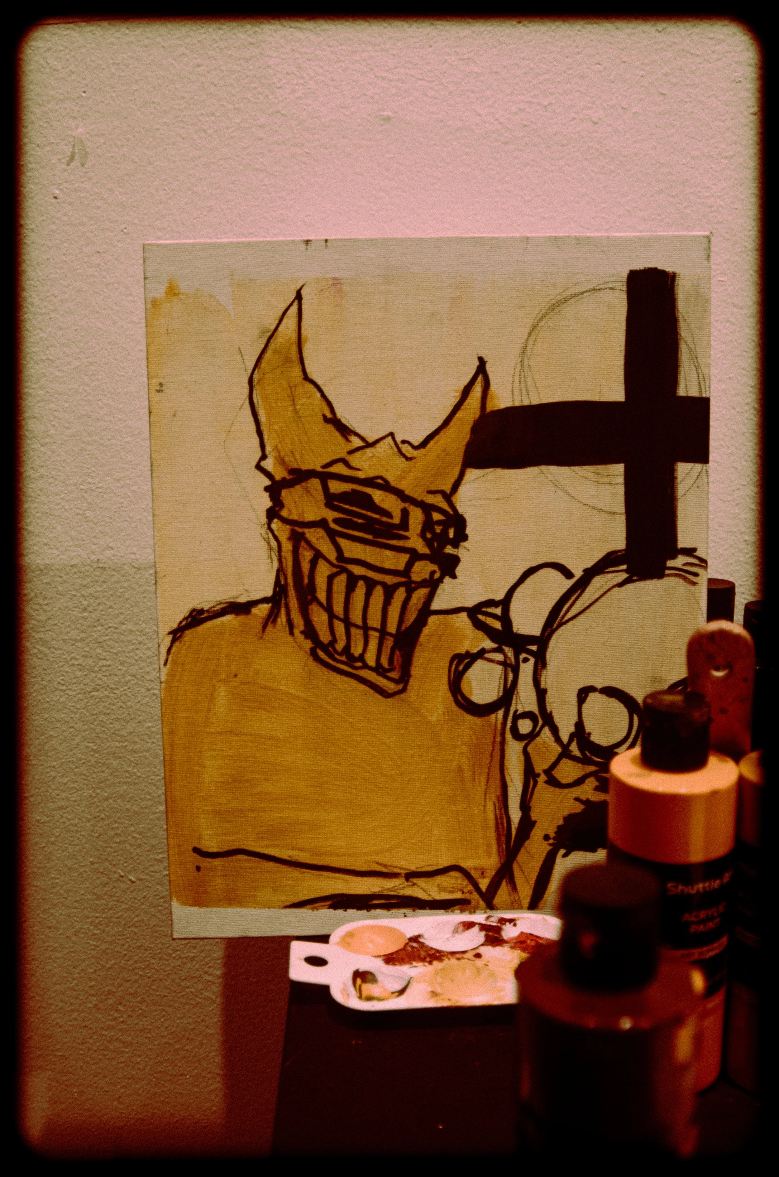 A line drawing of a spiky-haired character wearing glasses and a cape, with a large cross symbol on the right side. The drawing is on a piece of paper pinned to a wall, with art supplies in the foreground.