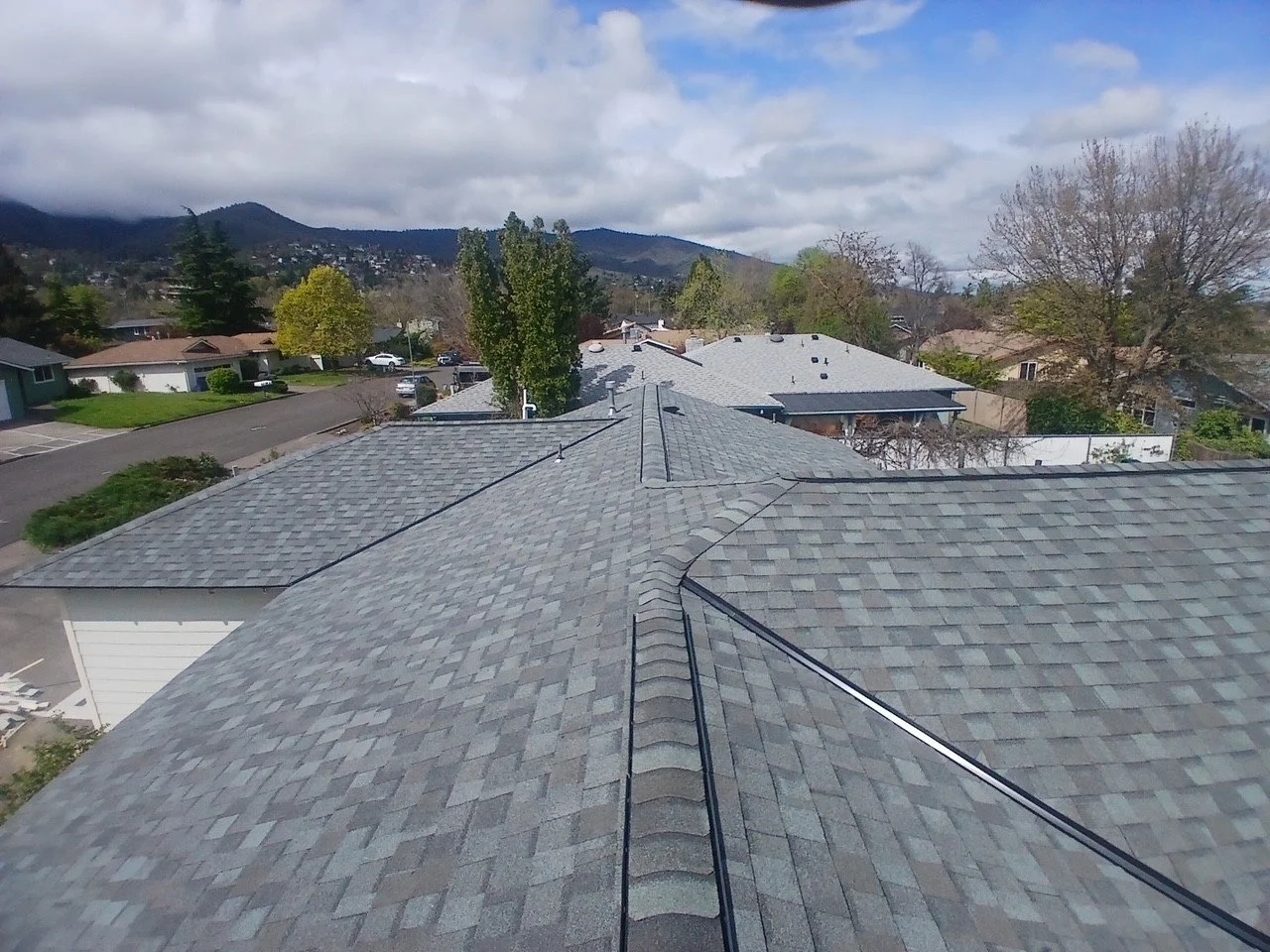 View of multiple rooftops with grey shingles, surrounded by trees and houses, with distant mountains and a partly cloudy sky in the background.