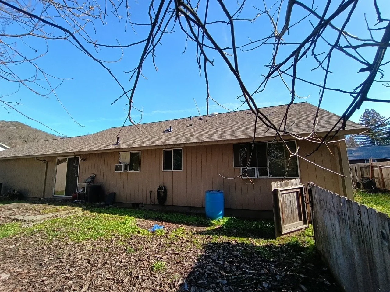 Backyard view of a single-story house with a brown roof, beige siding, and three windows. There are leafless tree branches in the foreground and a blue sky overhead.