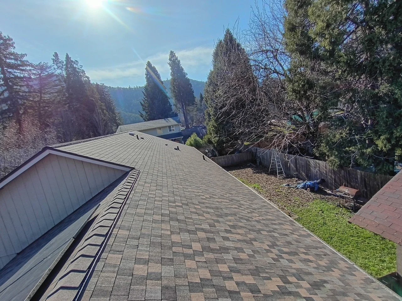 View of a house roof with shingles, a backyard with a fence, trees, and a mountain in the distance under a sunny sky.