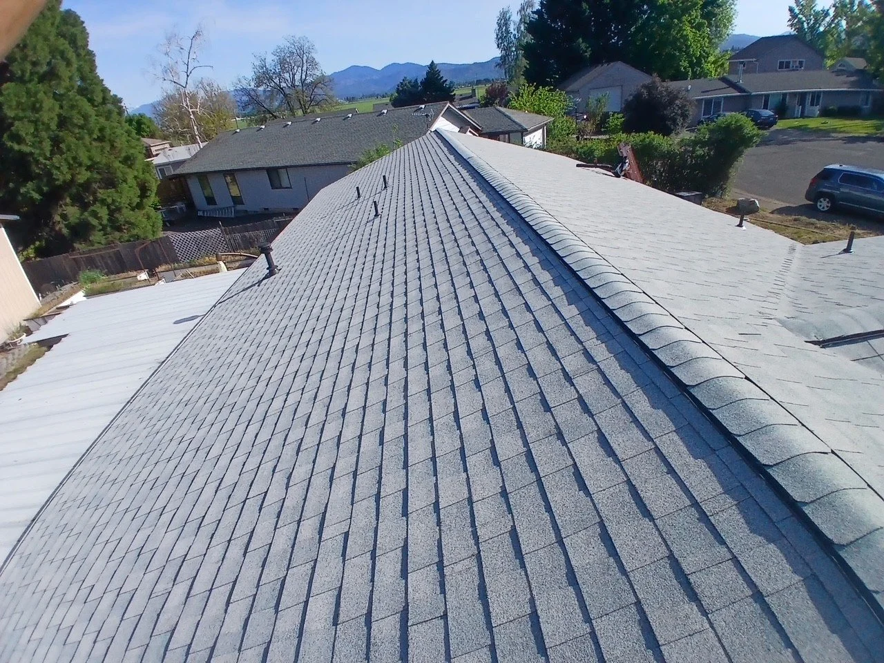 Aerial view of a house's gray shingled roof with several vent pipes and a ridge cap, surrounded by trees, other houses, and a parking lot with cars, mountain background, and clear sky.