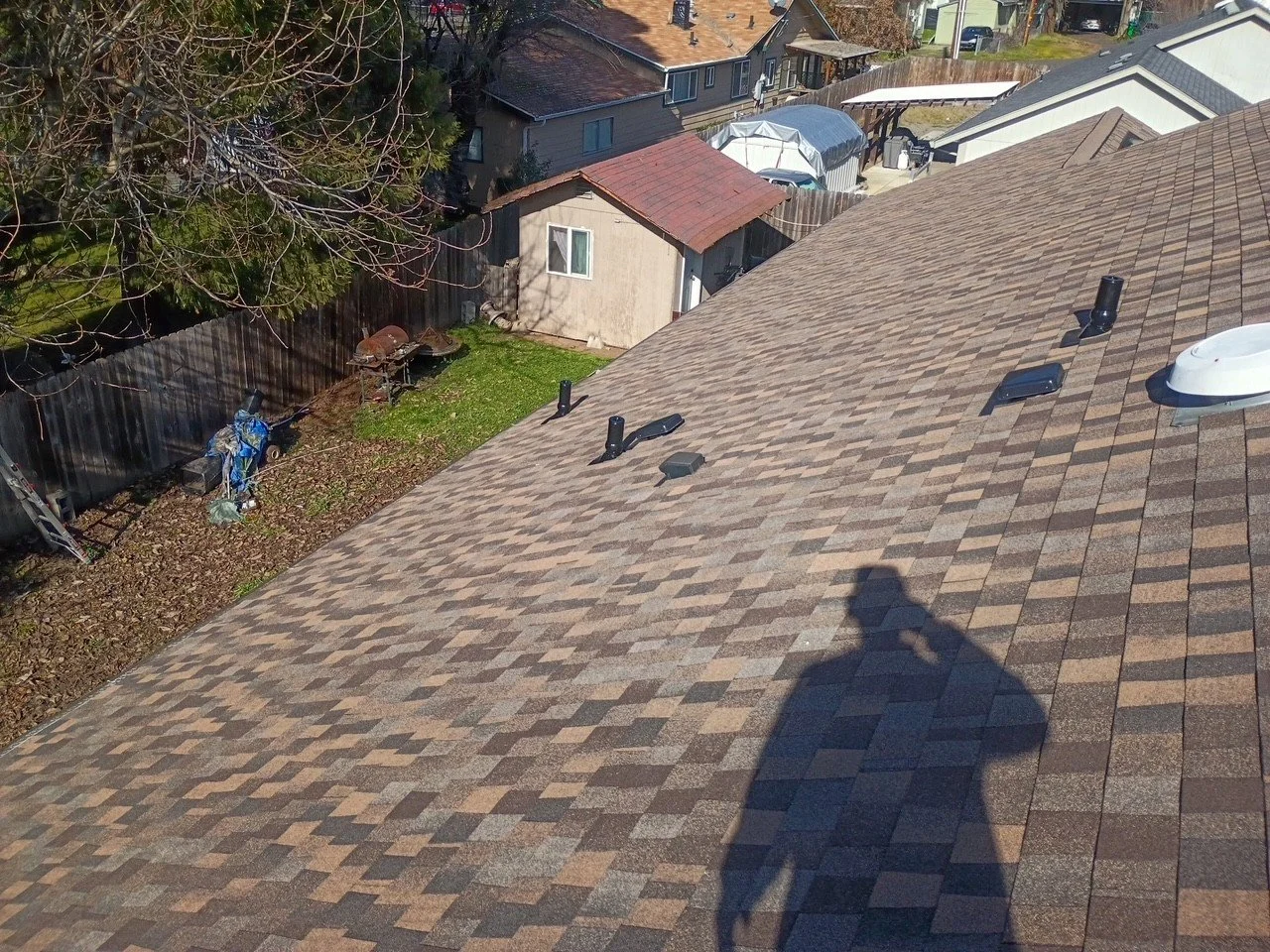 View of a residential rooftop with a shadow of a person taking a selfie. The roof has brown shingles with vents and a white attic vent. A backyard with a shed, trees, and a small garden is visible below.