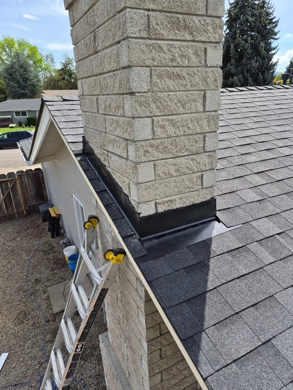 Close-up of a brick chimney on a residential roof with shingled roofing, construction tools, and a ladder nearby, indicating ongoing roofing or chimney repair.