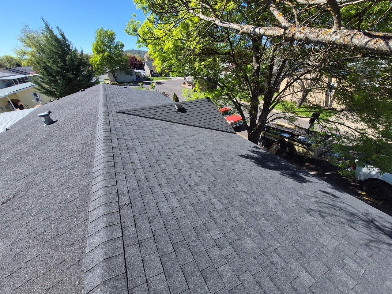 Aerial view of a house roof with gray shingles, a vent pipe, and surrounding trees and neighborhood street with parked cars.