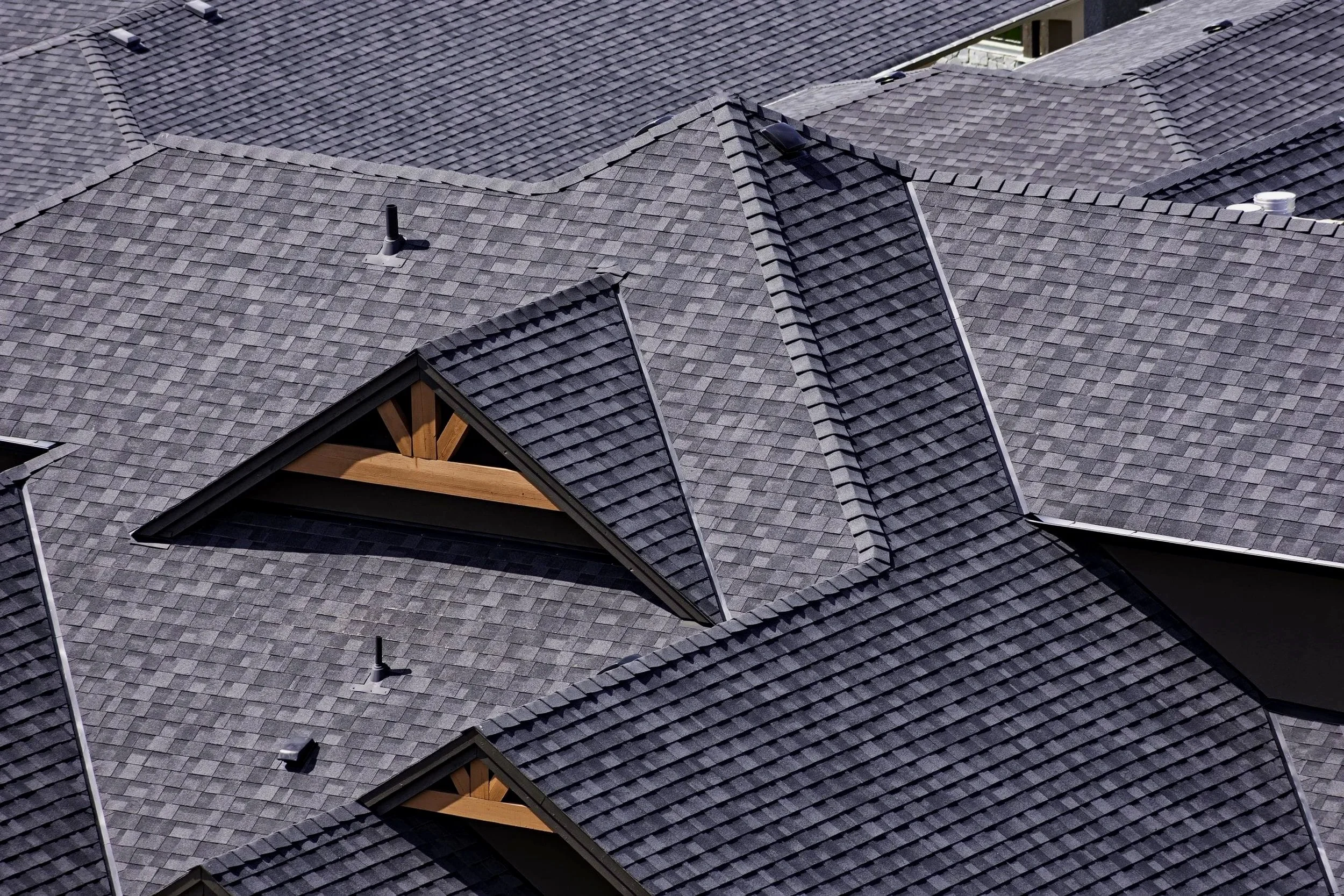 An aerial view of a residential roof with multiple intersecting sections covered in gray asphalt shingles, including small vents, pipes, and dormer windows.