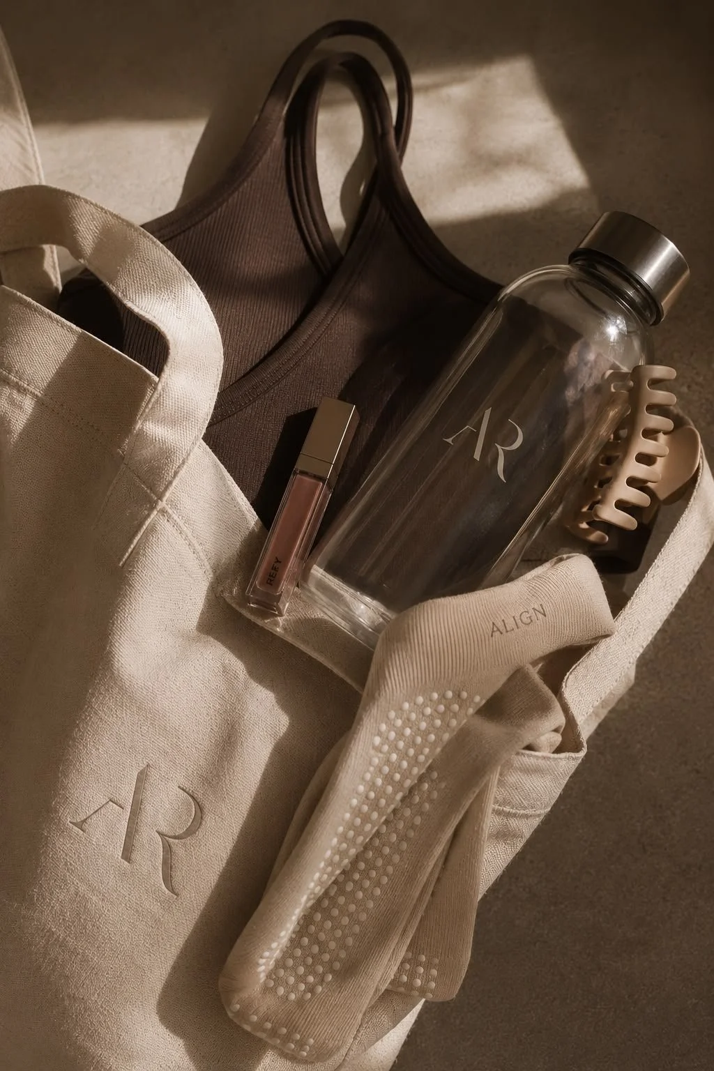 A tote bag contains a brown tank top, a white tote bag, a clear water bottle with the word 'AR' on it, a lipstick, a hair claw clip, and a beige sweatband with the word 'ALIGN' on it.