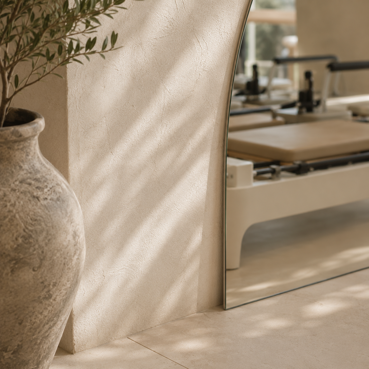 A marble floor beside a textured beige wall with a potted olive tree, and a large rectangular mirror reflecting a medical or therapy room with a padded examination table.