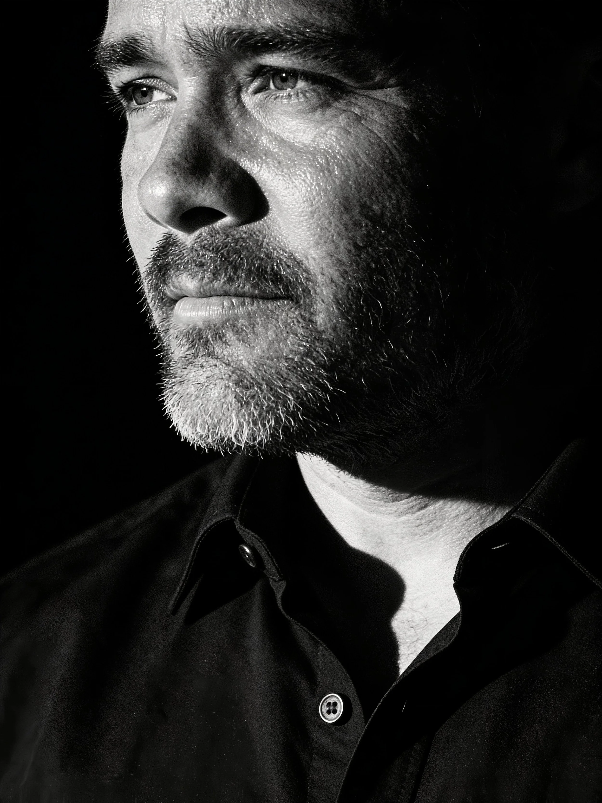A black and white close-up portrait of a man with light skin, showing his face and part of his upper body. He has short hair, a beard, and is wearing a dark shirt with buttons. The lighting emphasizes the texture of his skin and facial hair, against a dark background.