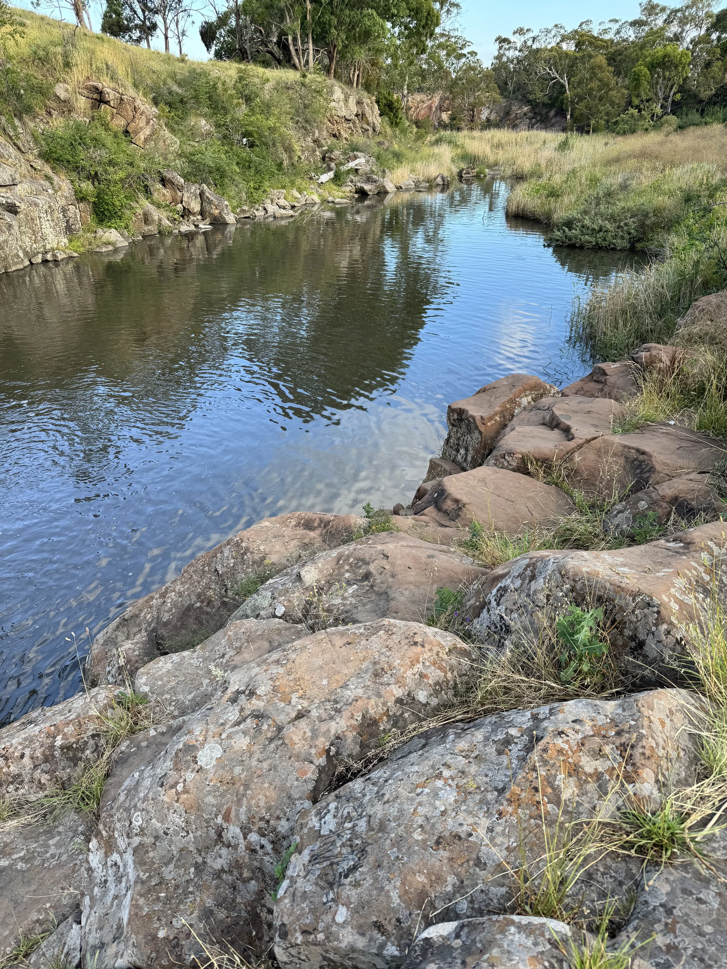 A peaceful river scene with rocks along the riverbank, green trees and grass on the hillside, and a clear sky reflected in the water.
