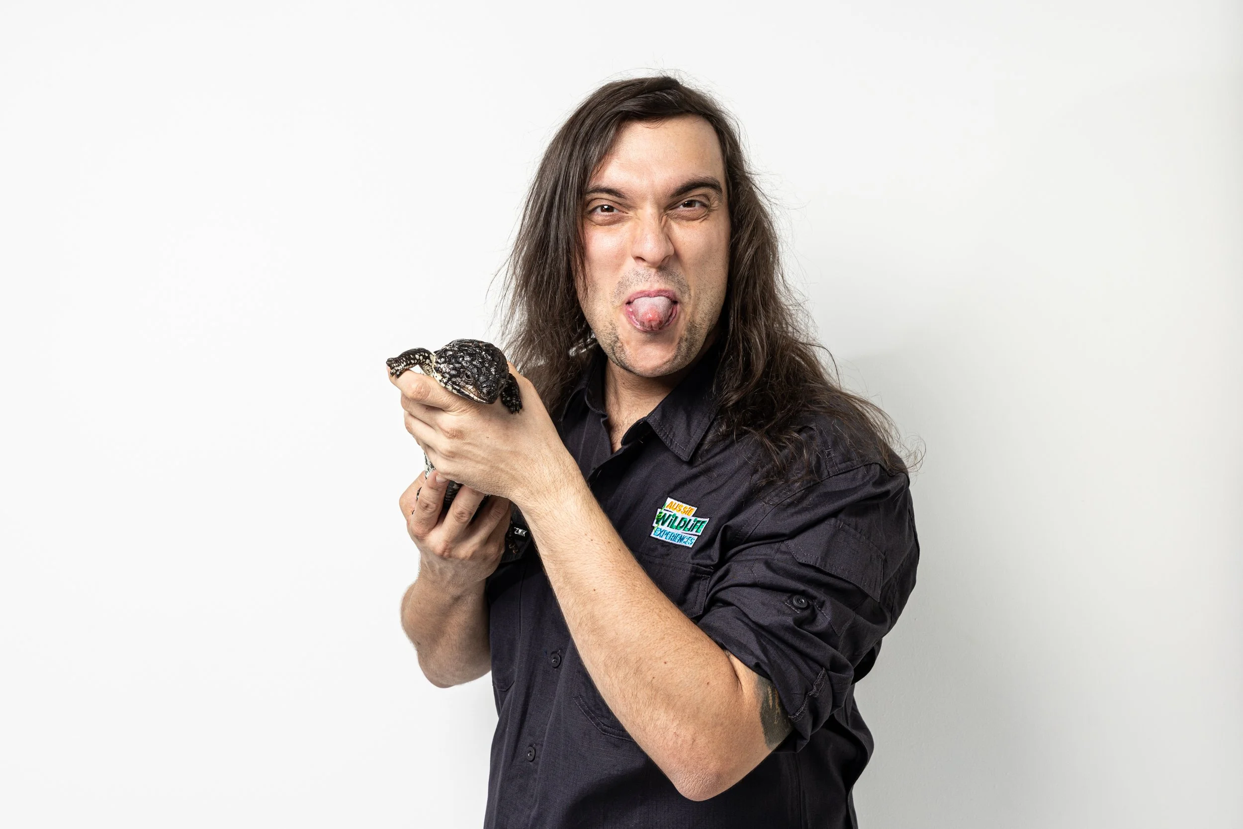 A man with long dark hair making a playful face with his tongue out and holding a black and white Shingleback Lizard in his hand. He is wearing a dark shirt with a colourful patch.