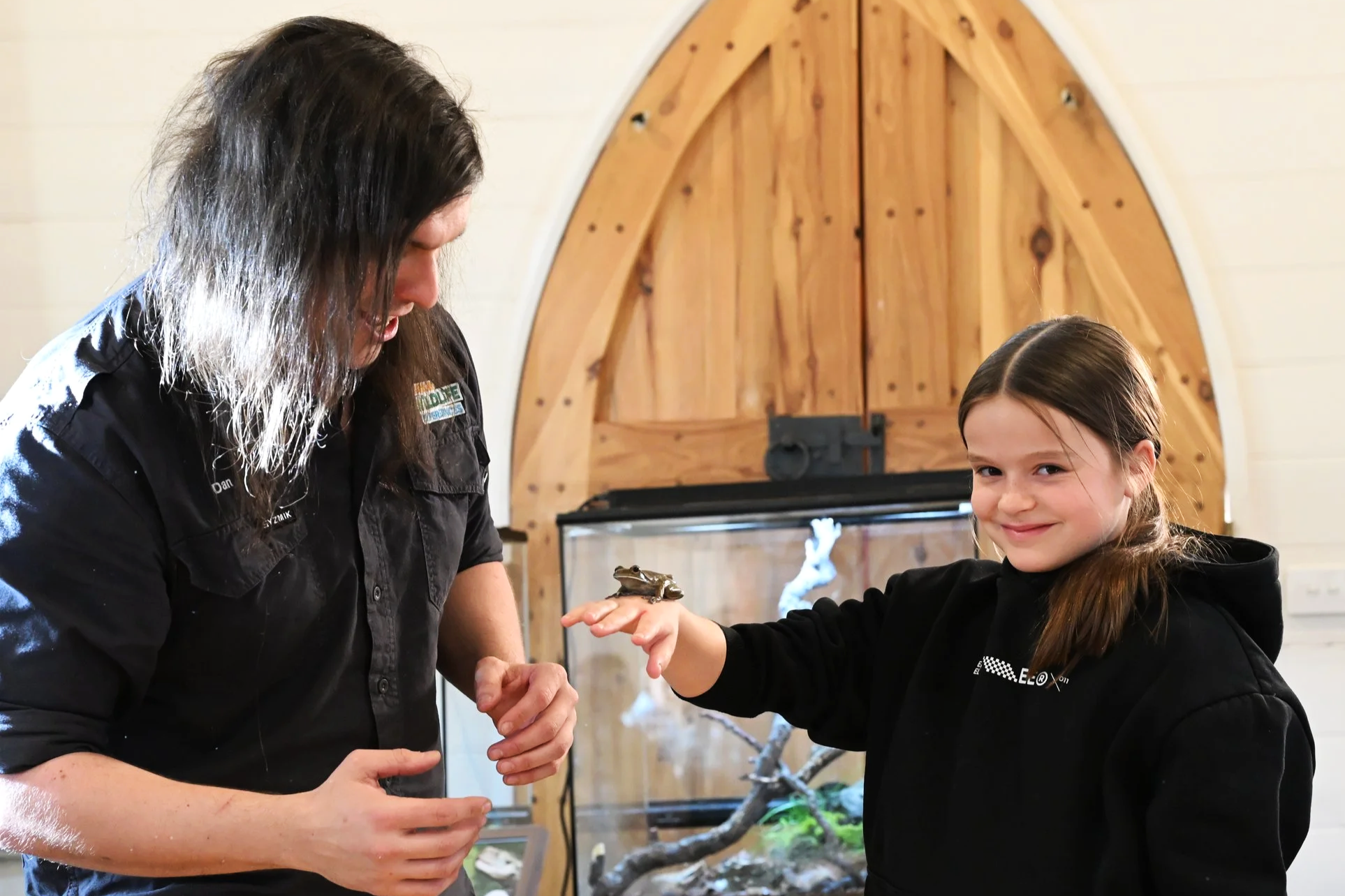 A young girl with long brown hair and a black hoodie smiling as she extends her hand to a small frog perched on her palm, with a man with long dark hair looking on