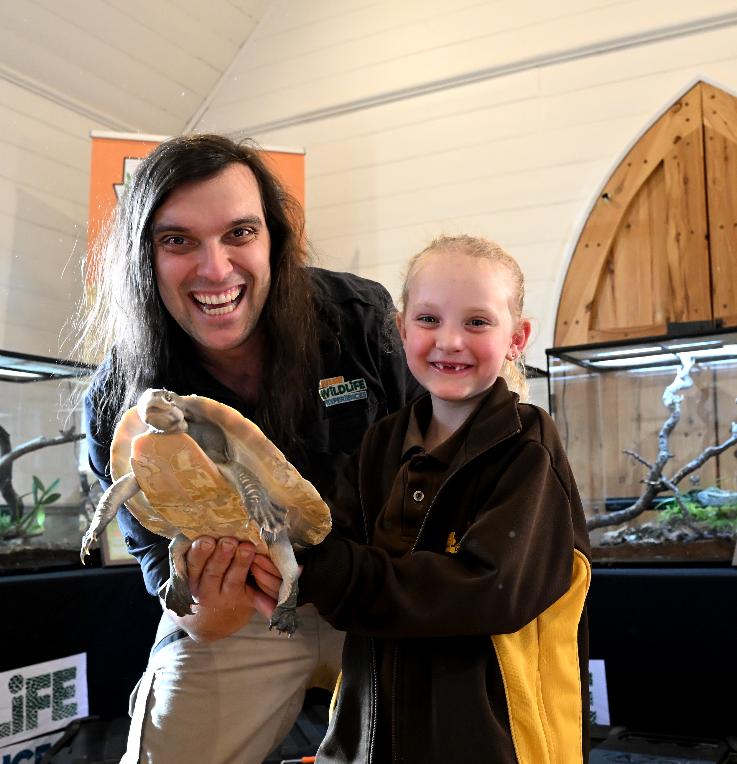 A man and a young girl smiling while holding a turtle at a wildlife exhibit with wooden and metal structures in the background.